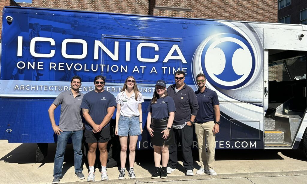 Six Iconica team members stand smiling in front of a large blue company truck with Iconica branding and the slogan “One Revolution at a Time” on a sunny day.