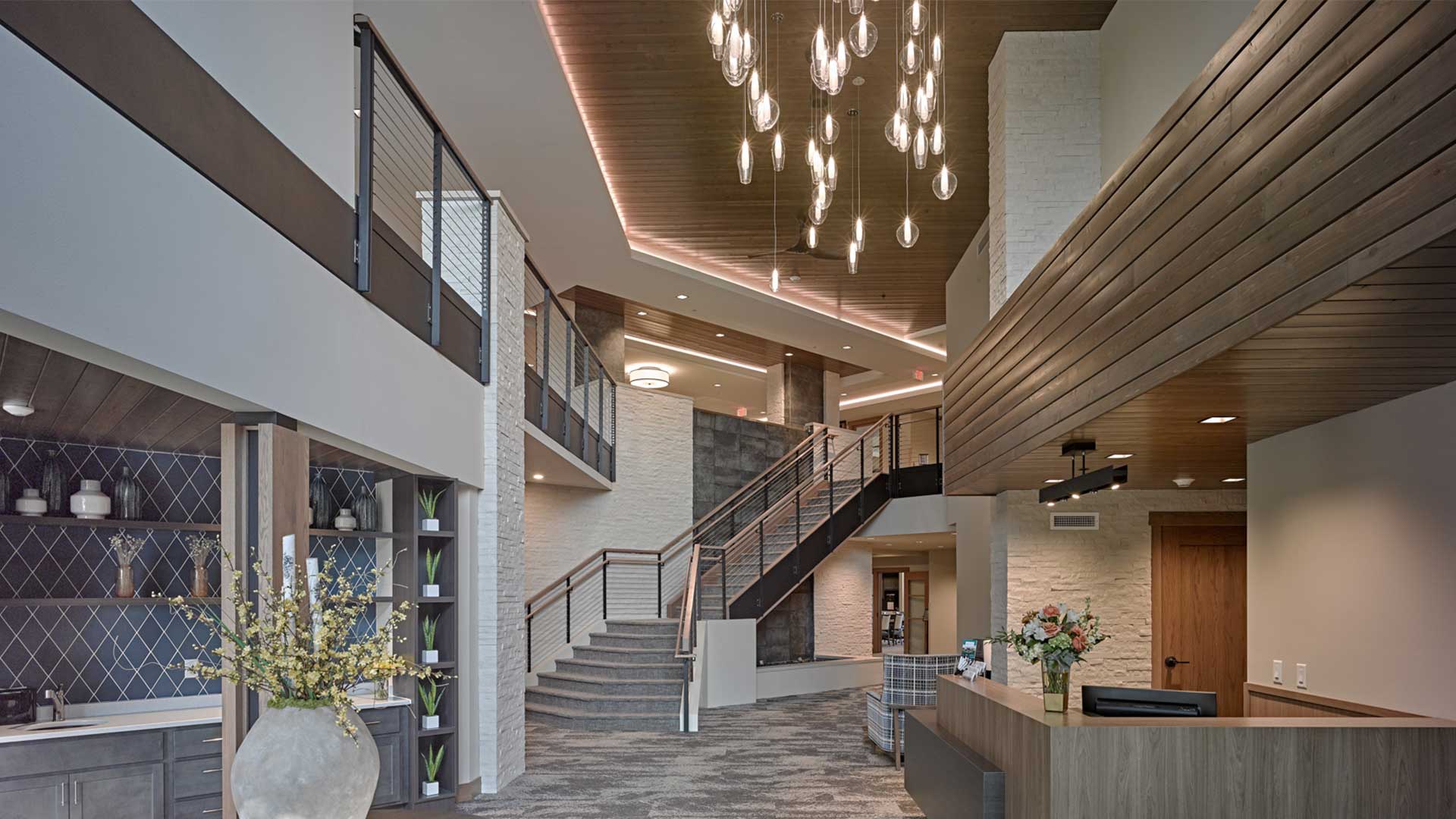 Modern senior living community lobby with reception desk, staircase to second-floor mezzanine, warm wood ceiling, and hanging pendant lights.