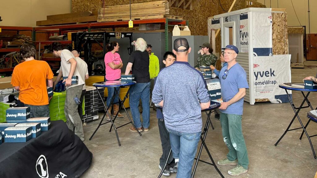 Students And Iconica Team Members Gathered Around High-top Tables In A Workshop Space, Eating Jersey Mike's Subs During A Lunch And Learn Event, With Lumber Racks, A Forklift, And Tyvek HomeWrap Materials Visible In The Background.