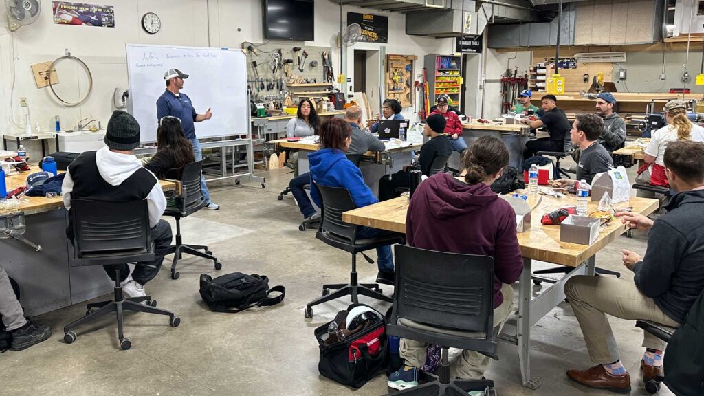 An Iconica Presenter Stands At A Whiteboard Speaking To MATC Students Seated At Workshop Tables Eating Lunch, In A Carpentry Shop Filled With Tools And Equipment.