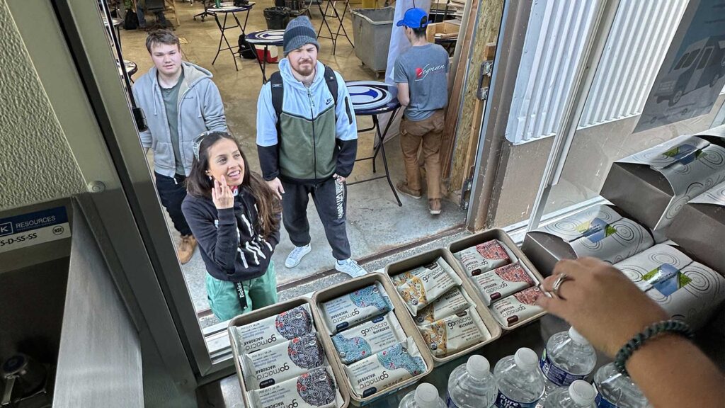 View From Inside The Iconica Food Truck Service Window Looking Out At Students Waiting To Order, With Trays Of GoMacro Protein Bars, Boxed Lunches, And Bottled Water Arranged On The Counter Ready To Be Served.