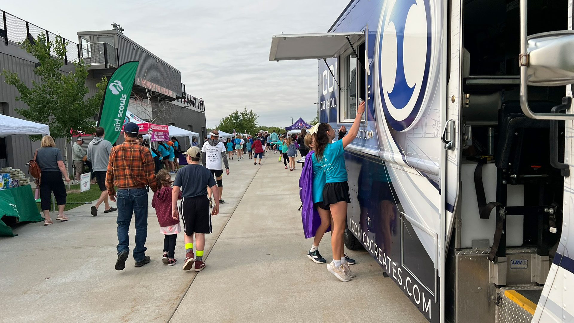 Community event with Iconica truck, vendor booths, and attendees in matching teal shirts walking along a paved path.