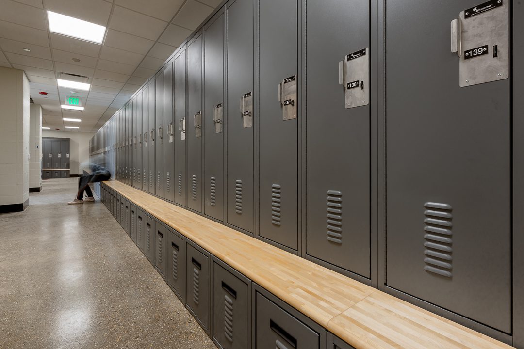 A close-up view of gray metal lockers with wood bench seating at the base, numbered in the 130s. A blurred figure crouches near the bench in the background, suggesting active use of the renovated locker room space.