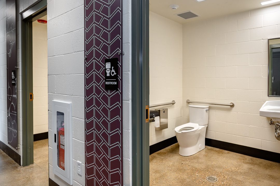 An accessible single-occupancy restroom with a wall-mounted toilet, grab bars, and a wall-hung sink. The door surround features a deep red decorative panel with a white geometric chevron pattern. An ADA-compliant restroom sign is mounted beside the door.