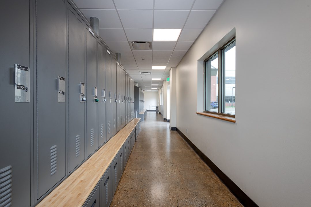 A bright locker room corridor with gray metal lockers and a wood bench running along the left wall. Windows on the right bring in natural light, with a view of an exterior building. The polished concrete floor reflects the overhead lighting.