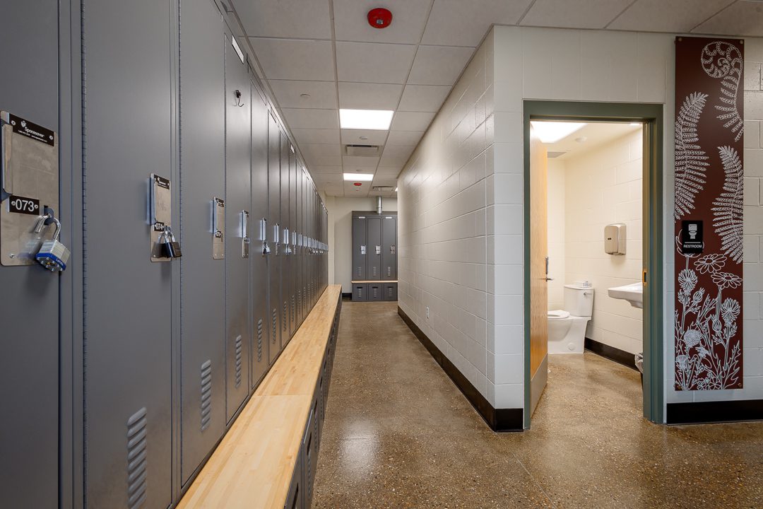 A locker room hallway with gray metal lockers and a wood bench on the left, leading to more lockers in the background. An open restroom door on the right reveals a toilet and sink. A deep red decorative panel with white botanical illustrations — ferns and wildflowers — lines the wall beside the restroom entrance.