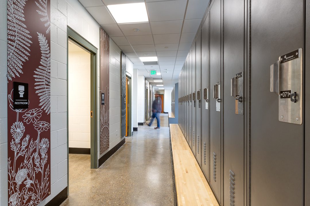 A locker room hallway with gray lockers and a wood bench on the right. On the left, a deep red wall panel with detailed white botanical illustrations flanks open restroom doors. A blurred figure walks through the corridor in the background.