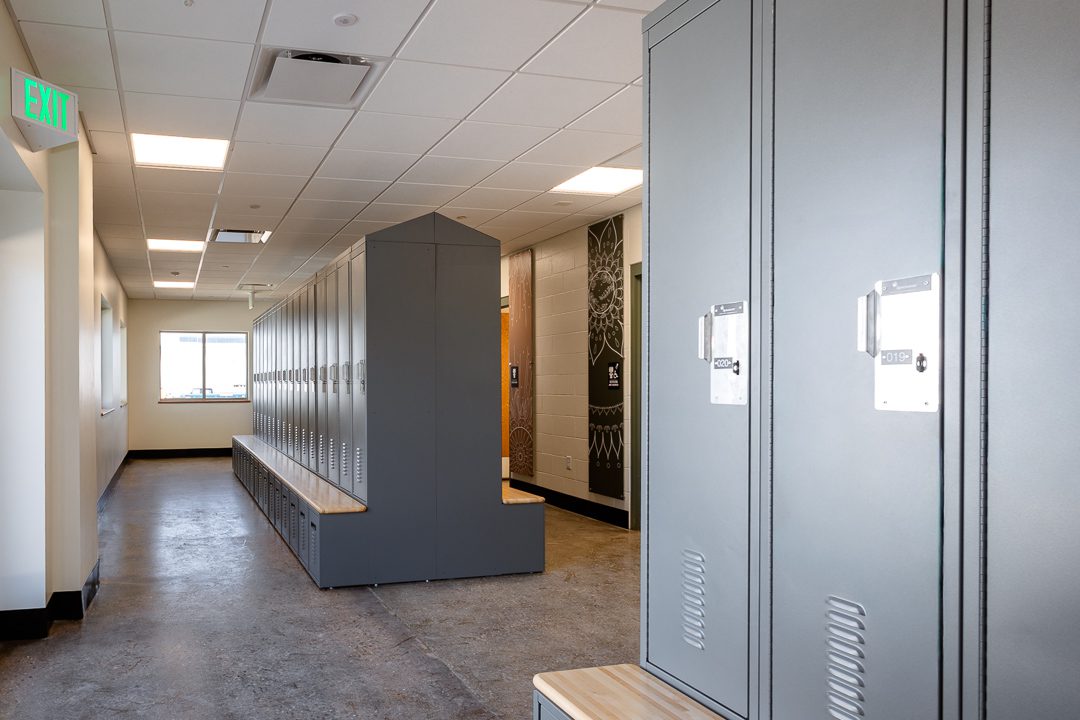A locker room with gray metal lockers and wood bench seating stretching down a long corridor. In the background, a decorative black wall panel with an intricate mandala-style illustration is visible beside a door surround. An exit sign is mounted near the ceiling.