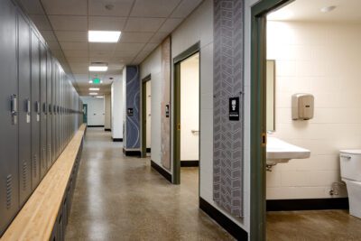 A Long Locker Room Corridor With Dark Gray Metal Lockers And Wood Bench Seating Along The Left Wall. On The Right, Multiple Restroom Doors Are Visible, With Decorative Wall Panels In Blue-gray Geometric And Floral Patterns Framing Each Entrance. The Hallway Extends Toward A Teal-colored Exit Door In The Distance.