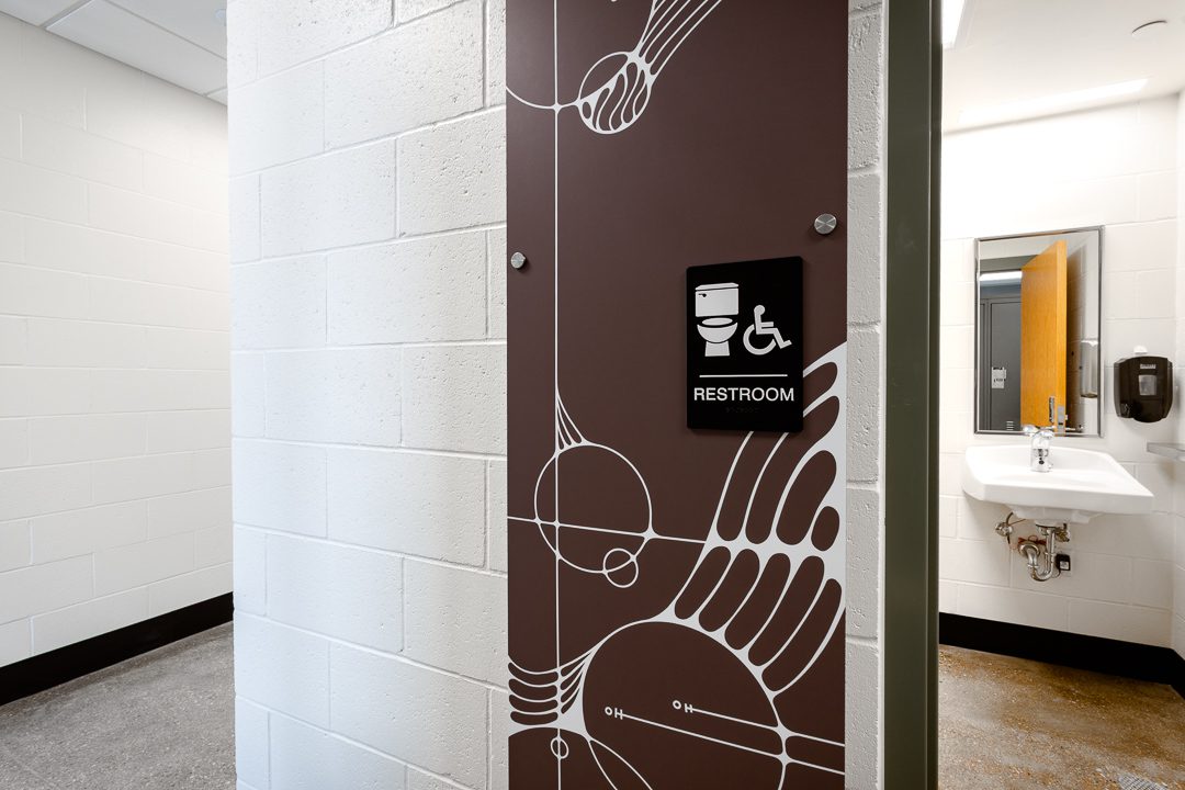 A close-up of a restroom entrance featuring a deep brown decorative wall panel with a white abstract bird illustration and molecular diagram motifs. An ADA-compliant restroom sign with wheelchair symbol is mounted to the panel. Inside, a wall-hung sink, mirror, and soap dispenser are visible.
