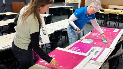 Two Iconica Volunteers Organize Pink Girls On The Run Materials And Directional Signs Across Tables Before An Event.