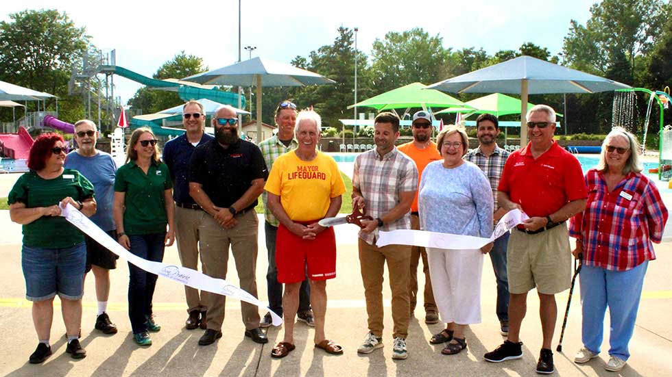 Community members and officials celebrate a ribbon-cutting ceremony at the newly renovated Sheehan Park Family Aquatic Center, with water slides and shade structures visible in the background