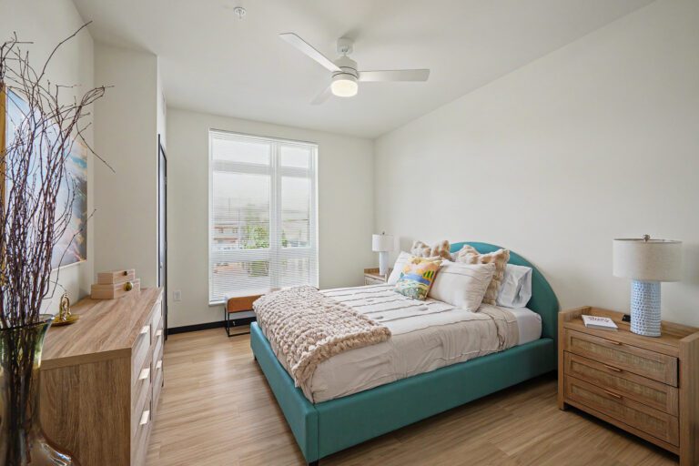 Vicino Apartments bedroom with teal upholstered bed, wood dresser, and large window with natural light