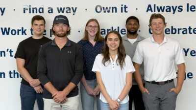 Group Photo Of Six Iconica Summer Interns Standing In Front Of A Wall Of Company Behaviors